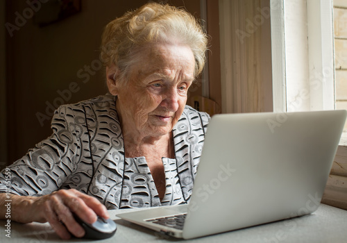 Obraz na płótnie An elderly woman sitting at the table and types on laptop. w salonie