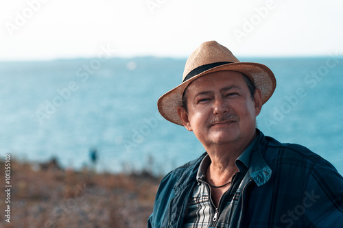 Adult smiling man with a mustache and beard in a straw hat against the sea. Close-up portrait of an elderly man. Sunny day, bright colors. Looking at the camera