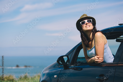 Relaxed happy woman on summer roadtrip travel vacation leaning out car window on blue sky background.