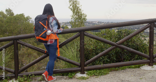 Young woman with a backpack standing with her back to the camera on a trail leaning on wooden rails admiring the mountain scenery