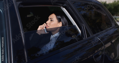Serious young female executive with button collar on phone and sitting in rear seat of limousine with window down