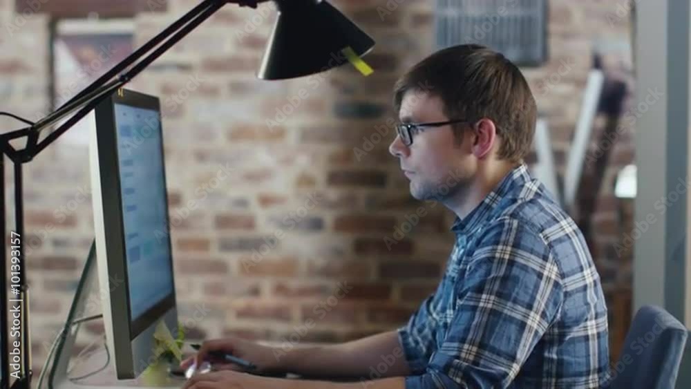 Young designer in glasses is sitting in front of a monitor in a loft and sketching on a graphic tablet. Shot on RED Cinema Camera.