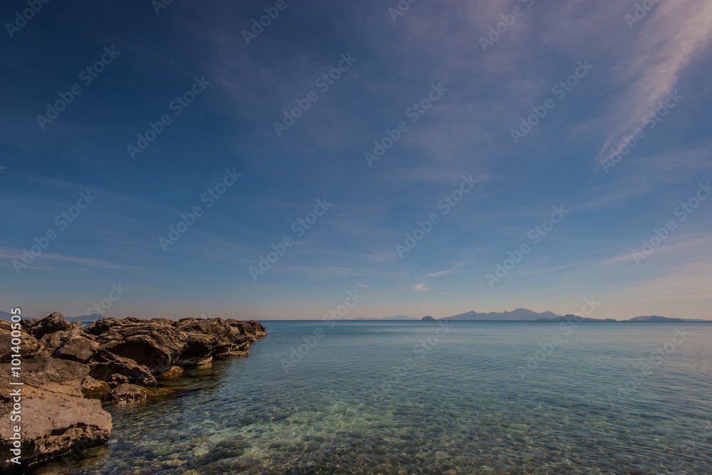 sea landscape of some rocks in the left hand side with a blue cloudy sky, water in the foreground and some islands in the background - obrazy, fototapety, plakaty 