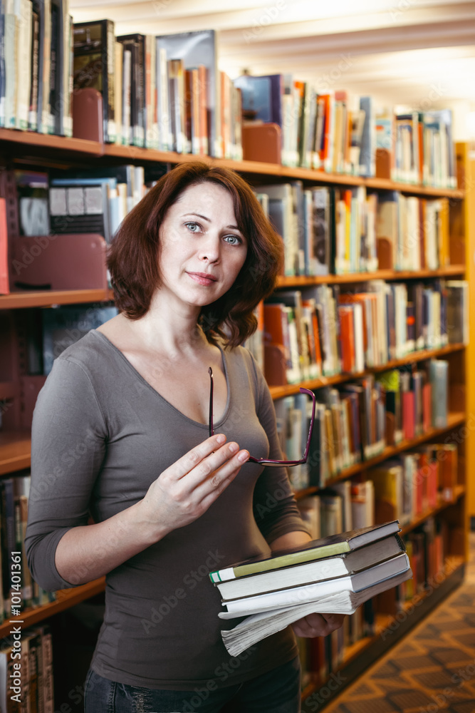 Portrait of middle age mature brunette Caucasian woman student with glasses in library holding book, looking directly in camera, teacher librarian profession, back to school concept Portrait of middle age mature brunette Caucasian woman student with glasses in library holding book, looking directly in camera, teacher librarian profession, back to school concept