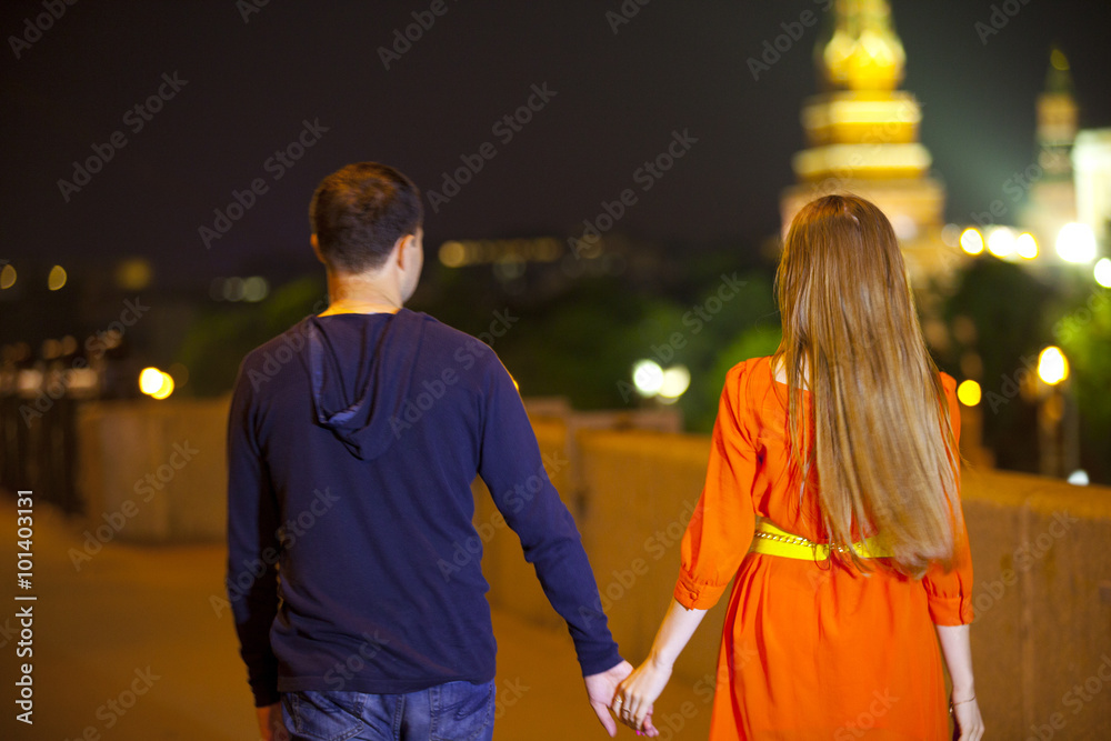 Пара - парень в синей футболке с капюшоном и девушка в рыжем платье с желтым поясом - гуляют по ночном городу