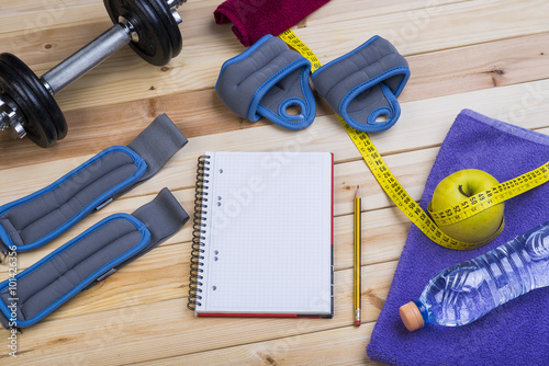 Sport Equipment. Dumbbells, Ankle Weights, Wrist Weights, Towel, Apple, Tape Measure, Bottle Of Water And Notebook To Workout Plan On Wooden Table. Sport Fitness Background