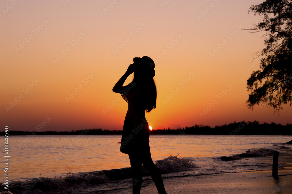 Silhouette of woman holding her hat, standing on the beach at sunset