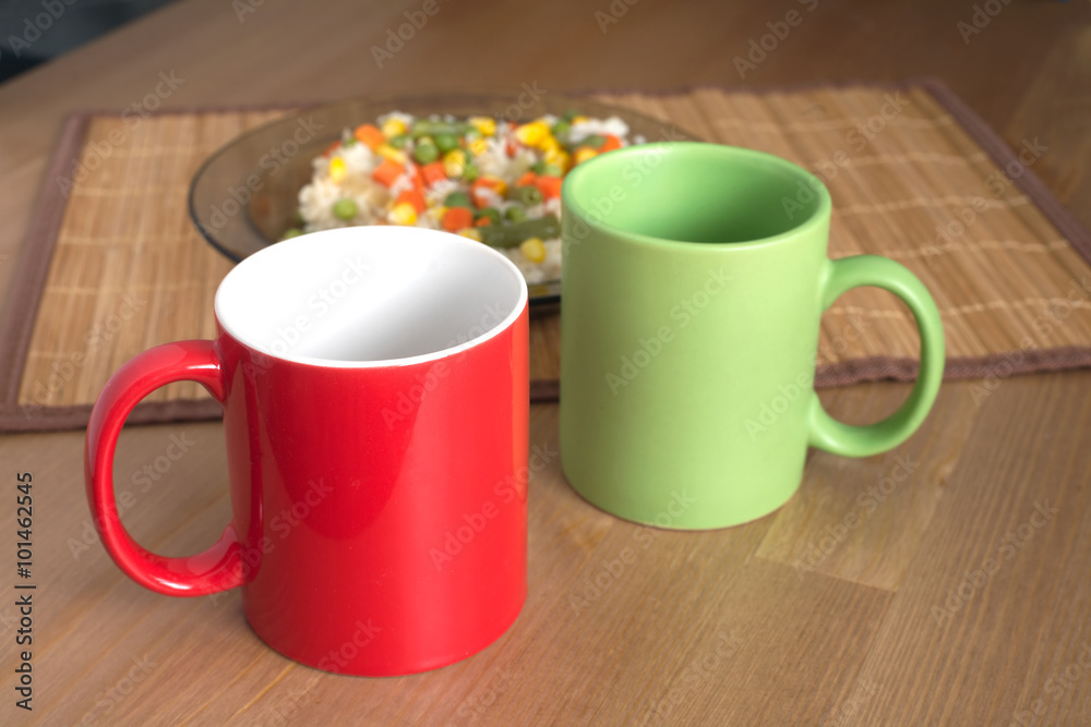 Red and green empty tea or coffee cups from ceramics and rice with vegetables on plate on brown wooden kitchen table closeup Red and green empty tea or coffee cups from ceramics and rice with vegetables on plate on brown wooden kitchen table closeup