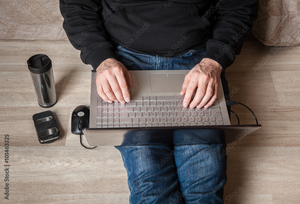 man sits on a wooden floor with a laptop on hir feet . the phone and a cup tumbler with coffee man sits on a wooden floor with a laptop on hir feet . the phone and a cup tumbler with coffee