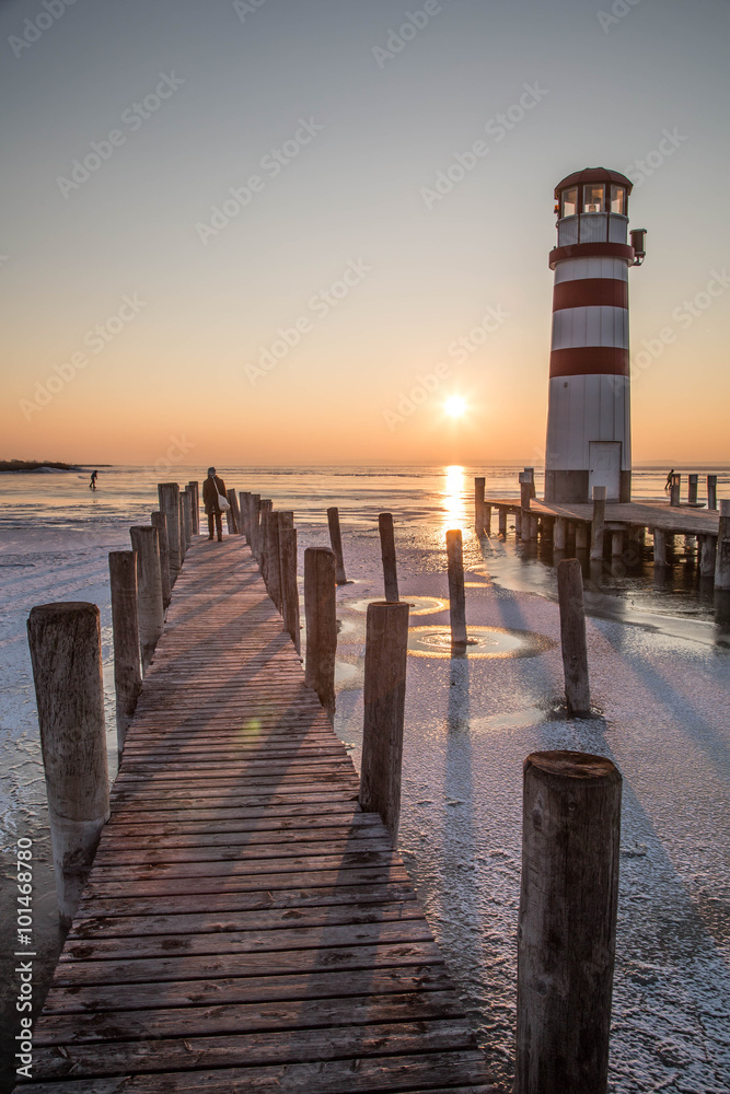 Holzsteg mit Leuchtturm im Winter vor gefrorenem See im Sonnenuntergang