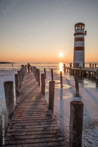 Holzsteg mit Leuchtturm im Winter vor gefrorenem See im Sonnenuntergang