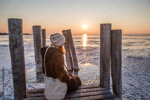 Frau sitzt am Holzsteg und blickt auf gefrorenen See bei Sonnenuntergang photo