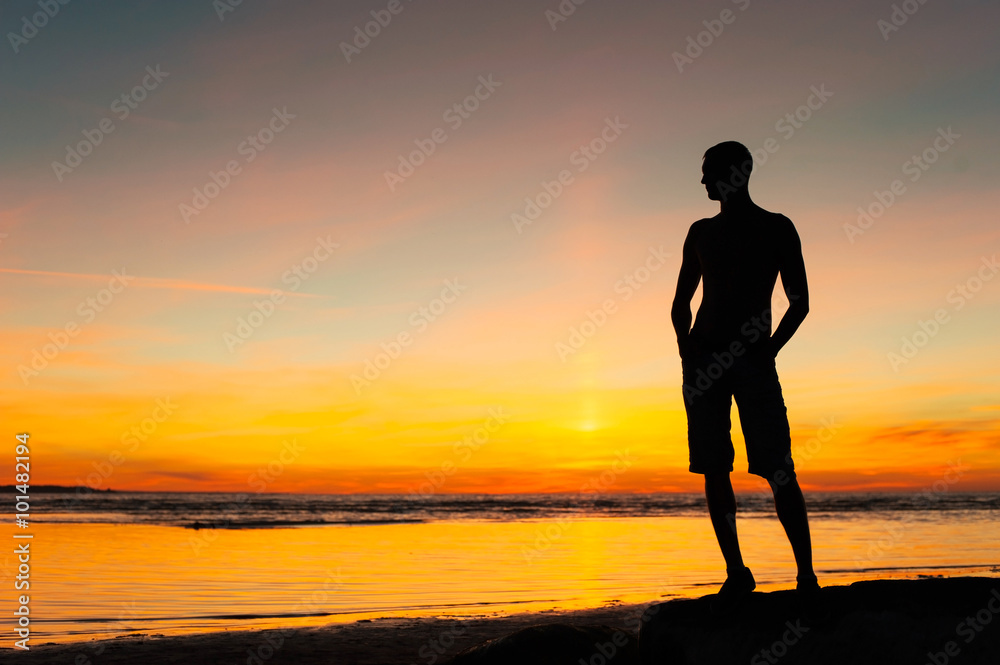 Young sporty man silhouette standing on the beach at sunset