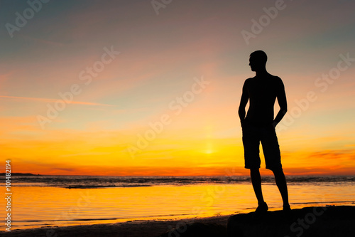 Young sporty man silhouette standing on the beach at sunset