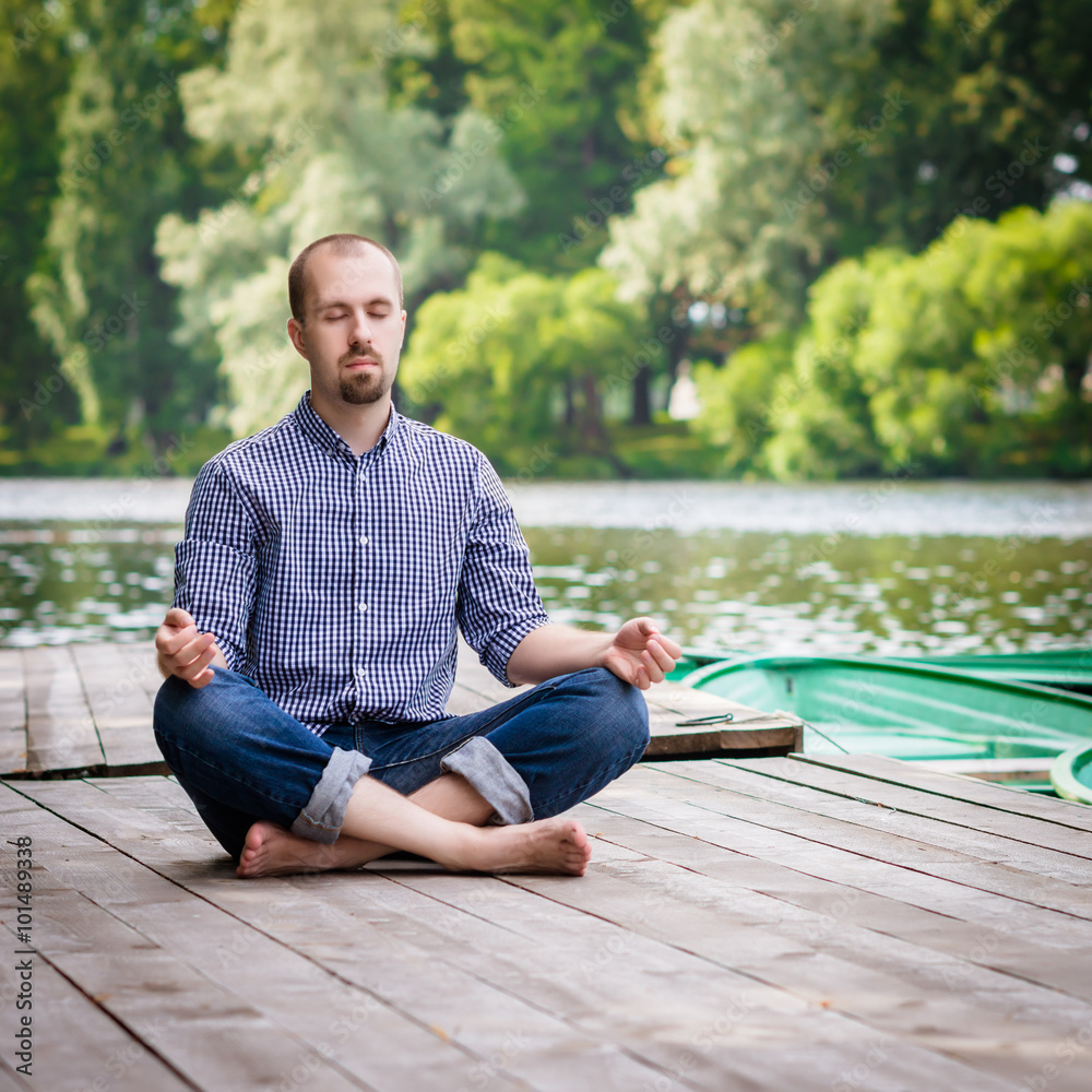 Obraz premium Young handsome bearded man sitting on wooden pier in summer day, meditating and relaxing