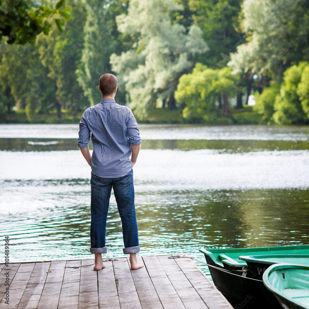 Young handsome man standing on wooden pier, relaxing and meditating in summer morning, back view
