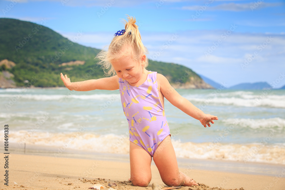 Naklejka premium Side View Little Blond Girl Sits on Sand at Shell by Surf