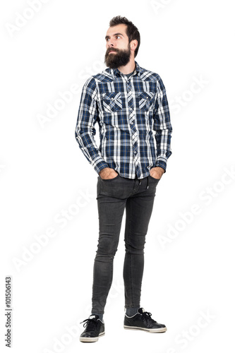 Serious bearded hipster in checked plaid shirt with hands in pockets looking away. Full body length portrait isolated over white studio background.