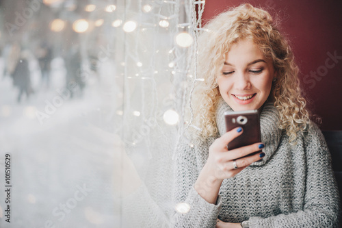 Young blond curly female in warm sweater using a smartphone and smiling in cafe, winter city outside the window