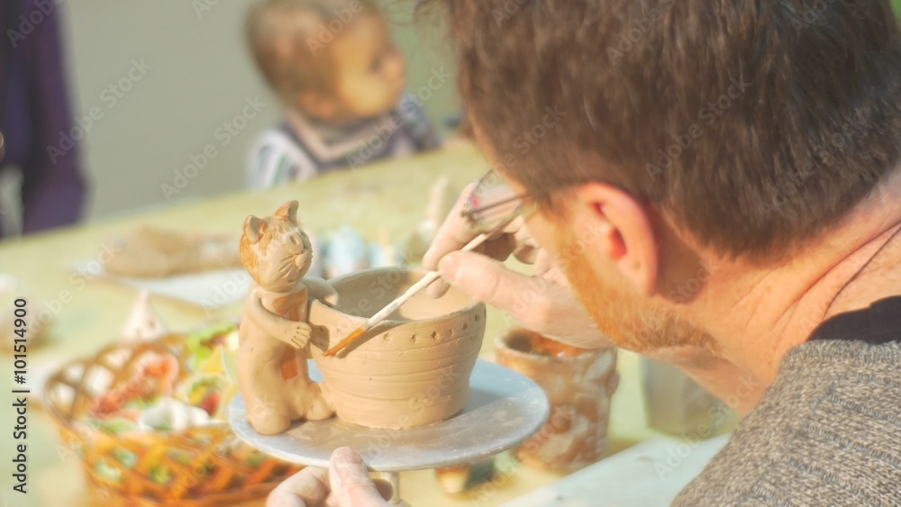 Man is Covering a Cat Statuette With Glaze by Brush Pot and Cat Teacher or Mom is Sitting with Kids Man in Glasses Learning a Pottery Students Workshop Man is Covering a Cat Statuette With Glaze by Brush Pot and Cat Teacher or Mom is Sitting with Kids Man in Glasses Learning a Pottery Students Workshop