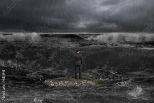 businessman standing on rock facing oncoming waves with dark oce