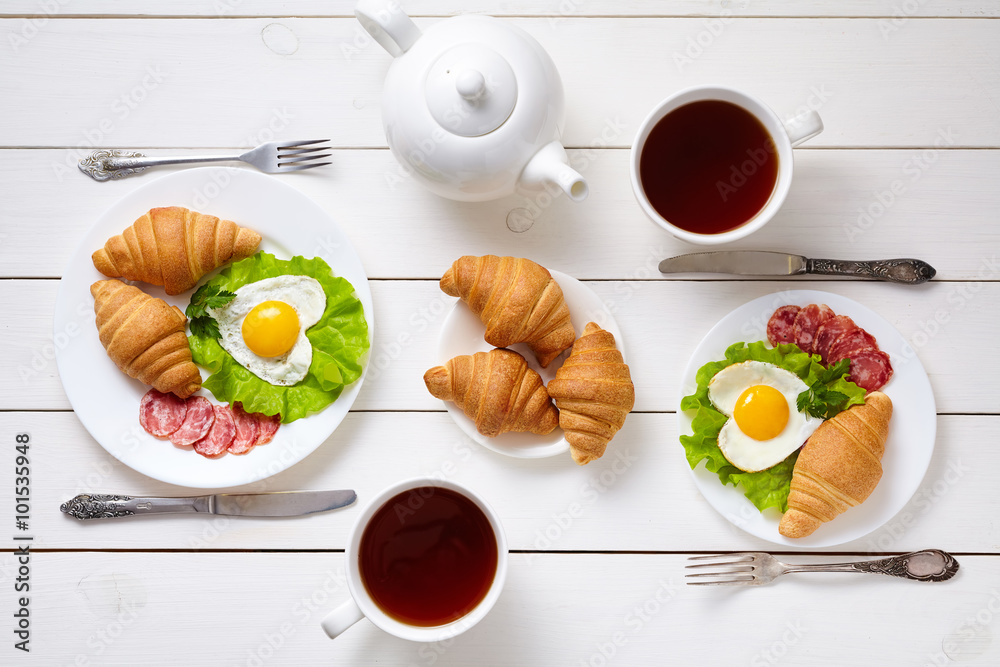 Φωτογραφία Heart shaped fried eggs, salad, croissants, salami sausage, composition and tea on white wooden table background