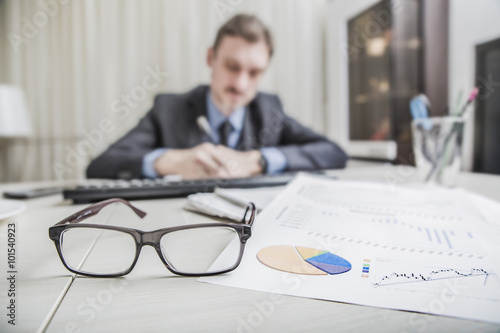 financial chart near dollars seen by unfocused glasses (only blurred silhouette of a businessman working at the Desk in the office visible viewed )