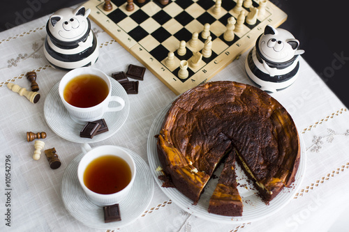 Шоколадный торт/брауни со сливочной начинкой, две чашки с чаем, шахматная доска и два керамических соусника в виде полосатых котов на скатерти
