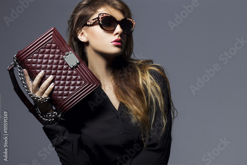portrait of a beautiful girl posing in studio in black shirt, blue jeans and fashion sunglasses, holding Burgundy handbag . The concept of stylish and sexy women. perfect skin and body photo