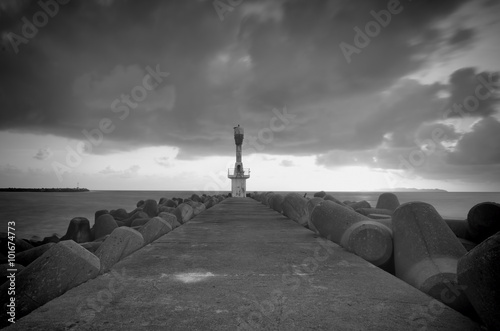 black and white image of lighthouse with dark and dramatic clouds during monsoon season.concrete breakwater and wall.