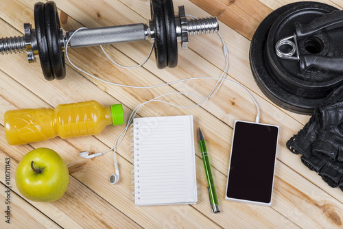 Sport Equipment. Dumbbells, Free Weights, Barbell, Hand Grip, Sport Gloves, Orange Juice, Smart Phone With Earphones And Notebook To Workout Plan On Wooden Table. Sport Fitness Background