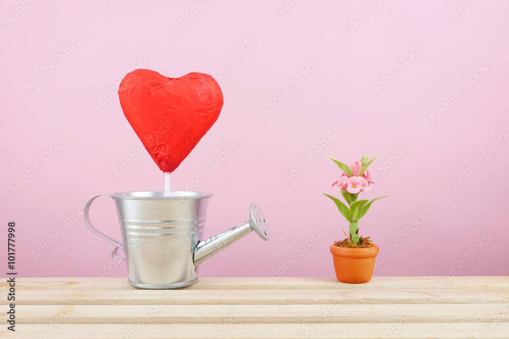 The red foiled chocolate heart stick with small silver watering can and mini fake flower in brown plant pot on wooden tray for Valentine's day. The red foiled chocolate heart stick with small silver watering can and mini fake flower in brown plant pot on wooden tray for Valentine's day.
