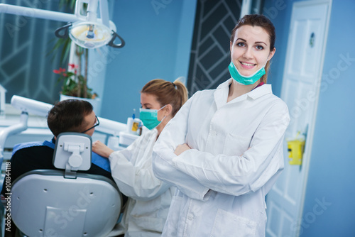 Portrait of beautiful dentist at office. Doctor and patient in background. Empty space for text.