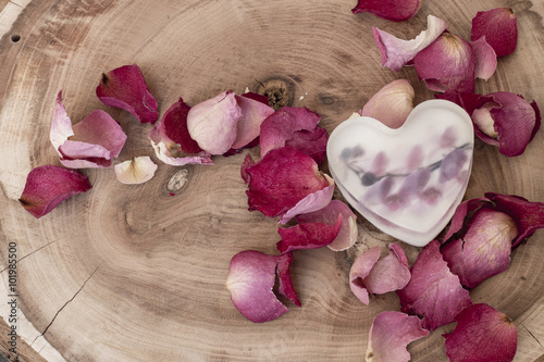 Soap in the shape of heart among rose petals on wood background 