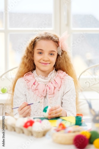 Beautiful young girl decorating Easter eggs at cozy home atmosphere.