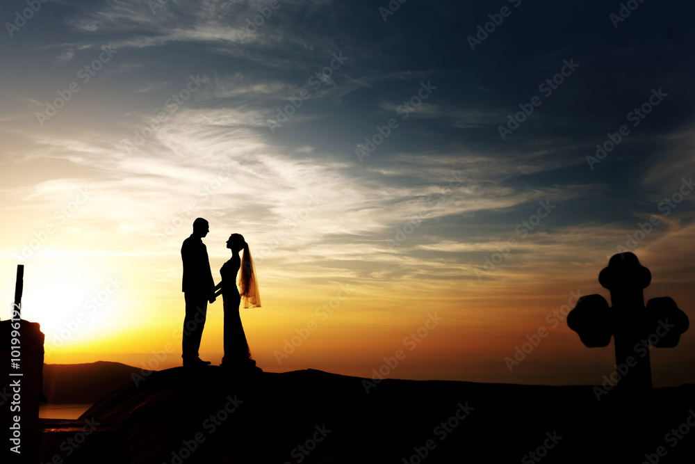 Groom and bride silhouette kissing on church roof at sunset sky