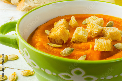 Pumpkin squash vegetable soup with croutons and pumpkin seeds in a green bowl on white wooden background, close up