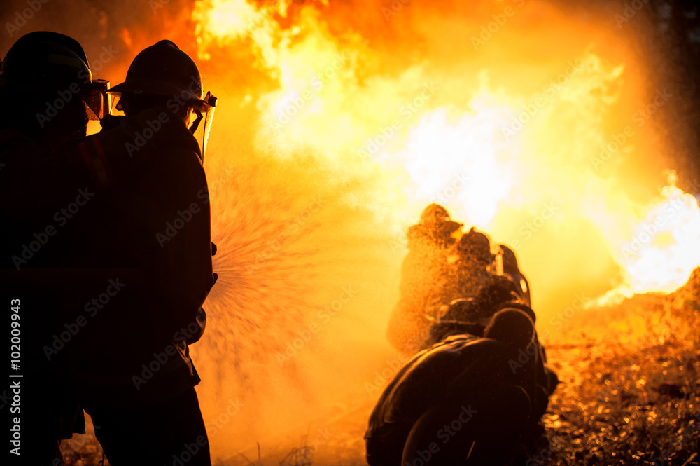 Fototapeta premium Silhouette of Firemen fighting a raging fire with huge flames of burning timber