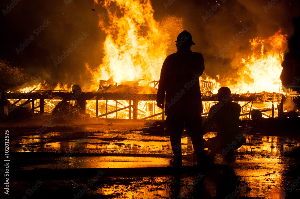 Fototapeta premium Silhouette of Firemen fighting a raging fire with huge flames of burning timber