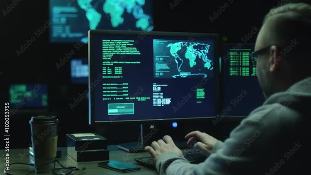 Male hacker in a hood works on a computer with maps and data on display screens in a dark office room. Shot on RED Cinema Camera.