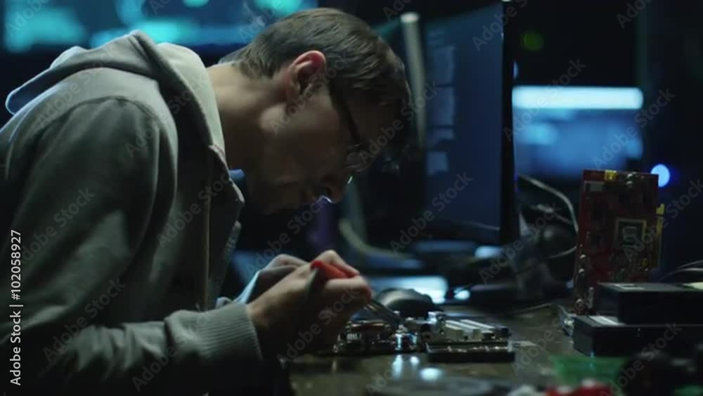 Electronics engineer is soldering an electric board with processors in a dark office with display screens. Shot on RED Cinema Camera. Electronics engineer is soldering an electric board with processors in a dark office with display screens. Shot on RED Cinema Camera.