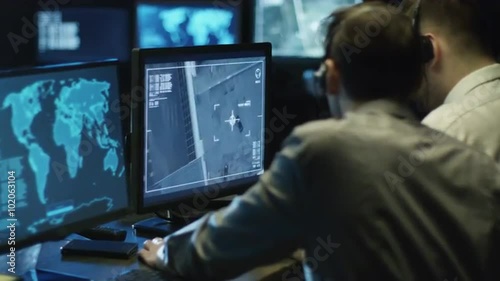 Two IT professionals are spying on businessman through satellite surveillance on computer in a dark monitoring room filled with display screens. Shot on RED Cinema Camera.
