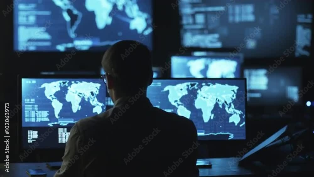 Male IT employee comes to monitoring room to work on computer with maps and data on display in a dark office room. Shot on RED Cinema Camera. Male IT employee comes to monitoring room to work on computer with maps and data on display in a dark office room. Shot on RED Cinema Camera.