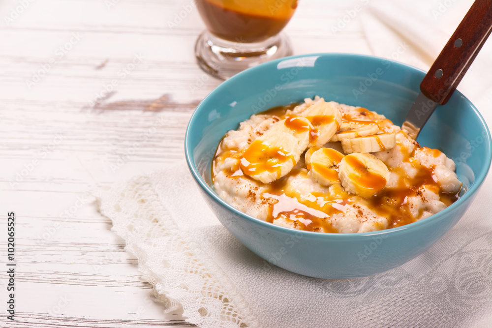 Oatmeal porridge with banana slices, cinnamon and caramel sauce over white wooden table. Selective focus Oatmeal porridge with banana slices, cinnamon and caramel sauce over white wooden table. Selective focus