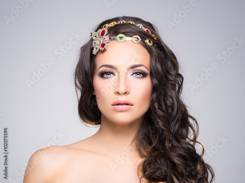 Attractive, curly brunette with flower alike golden headband with different gems over gray background.