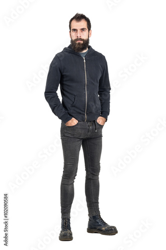 Tough confident stylish punker in black hooded sweatshirt looking at camera with hands in pockets. Full body length portrait isolated over white studio background.