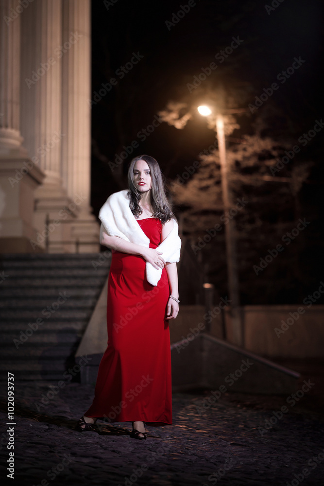Beautiful stylish girl with long hair, gorgeous makeup in a red evening dress with white fur collar is walking around the city in the summer one