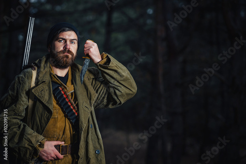 brutal hunter, bearded man in warm hat with a gun in his hand, a knife a backpack and smoking pipe in the dark wild forest in the autumn