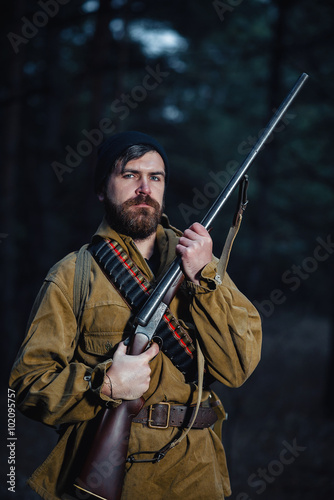 brutal hunter, bearded man in warm hat with a gun in his hand, a knife a backpack and smoking pipe in the dark wild forest in the autumn