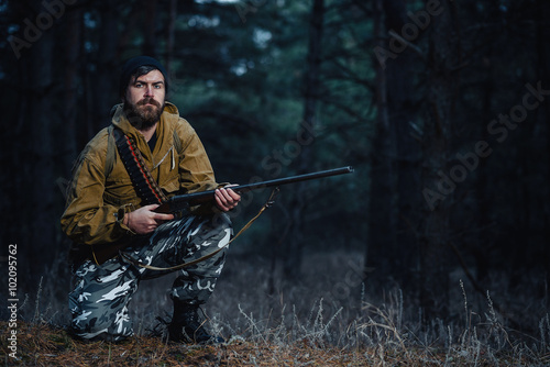 Photography brutal hunter, bearded man in warm hat with a gun in his hand, a knife a backpac